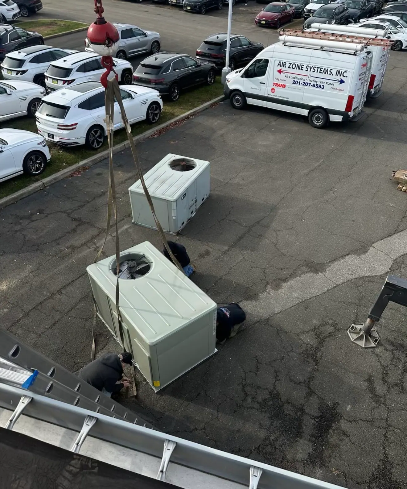 A packaged heating and cooling unit on lifting straps from a crane hook, with three workers beside it and two Air Zone Systems vans in the lot behind.