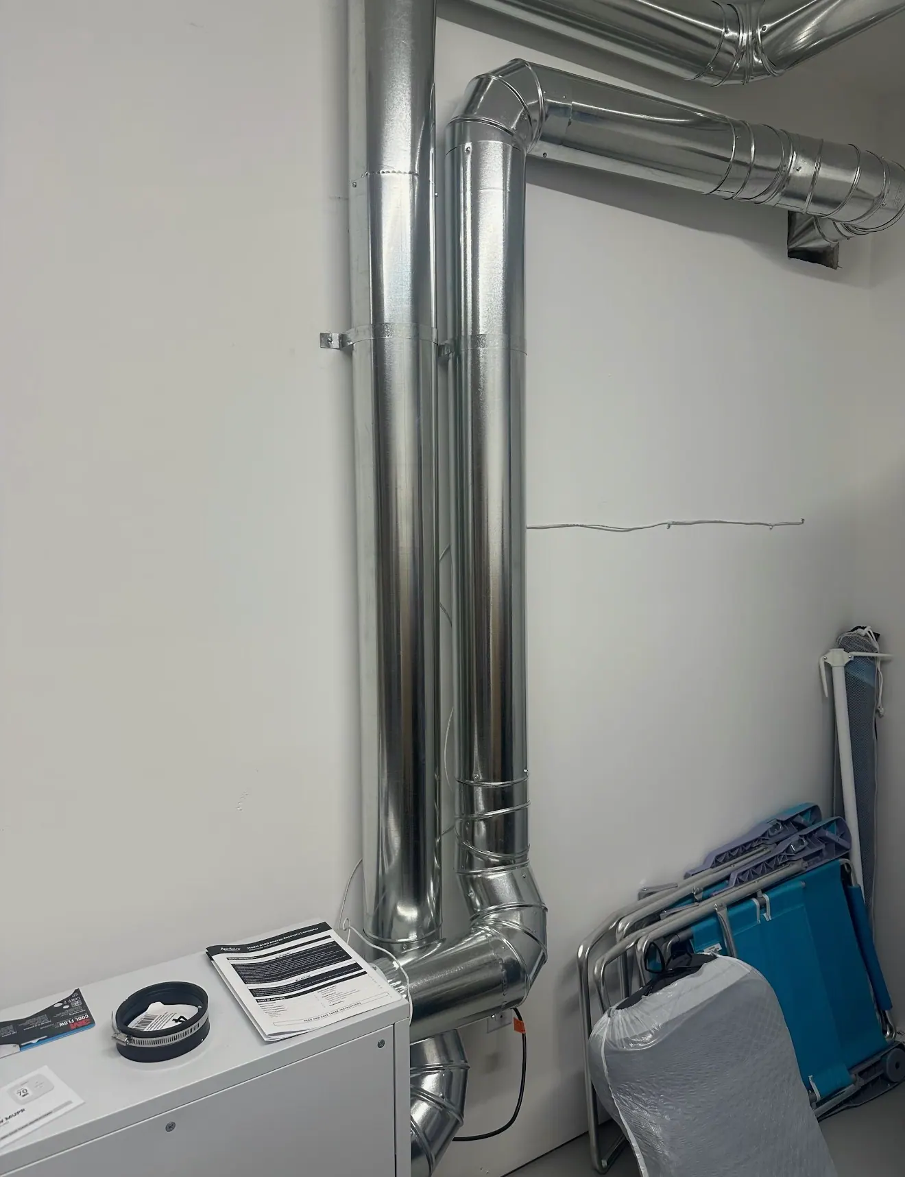 Two vertical galvanized round ducts running down a white wall to a white AprilAire unit at the lower left.