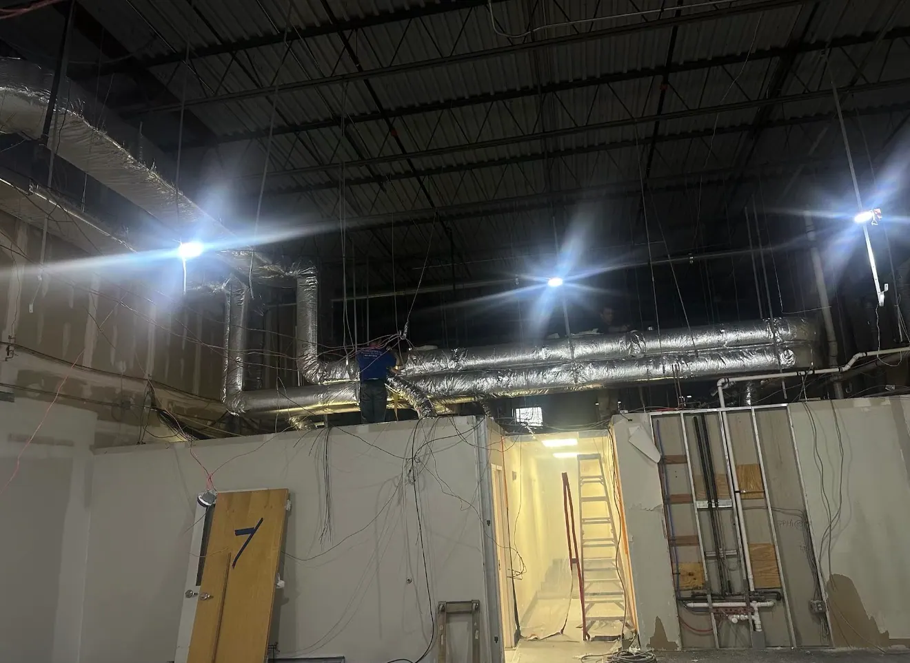 Insulated overhead ductwork in a large unfinished building, with a worker at a duct junction above a partition wall.